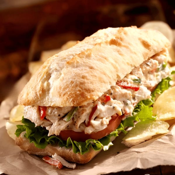 Chicken salad sandwich on a baguette with lettuce, tomato, and potato chips on parchment paper.