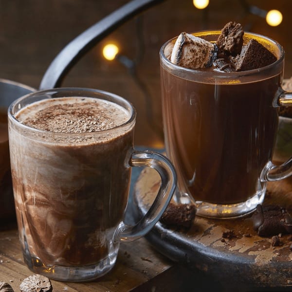 Two mugs of hot chocolate, one with marshmallows and brownie pieces, on a wooden surface.