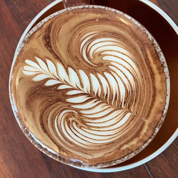Close-up of a latte in a brown mug with intricate leaf-shaped latte art on a wooden table.