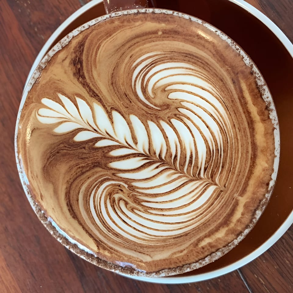 Close-up of a latte in a brown mug with intricate leaf-shaped latte art on a wooden table.