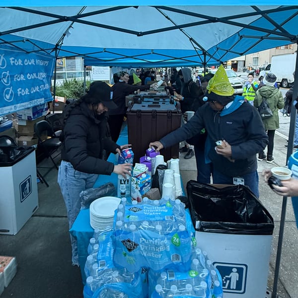 People under a blue tent serving drinks and snacks from a table with bottled water and milk cartons.