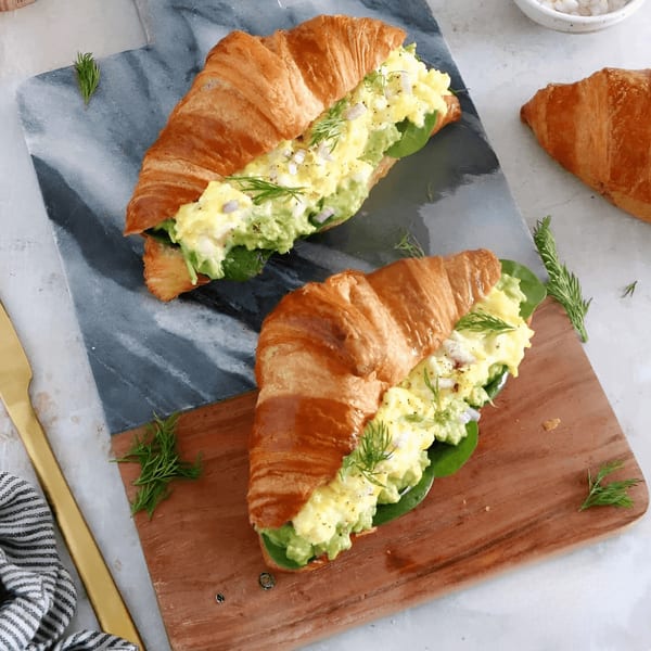 Two croissants filled with egg salad, avocado, and spinach, garnished with dill.