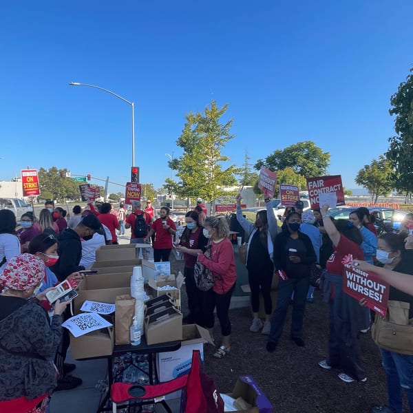 Nurses and healthcare workers in red shirts protest with signs reading "On Strike" and "Fair Contract Now".
