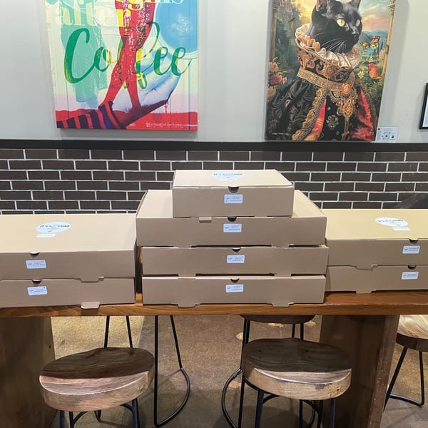 Numerous "It's a Grind" coffee boxes stacked on a wooden table with stools, under vibrant art.