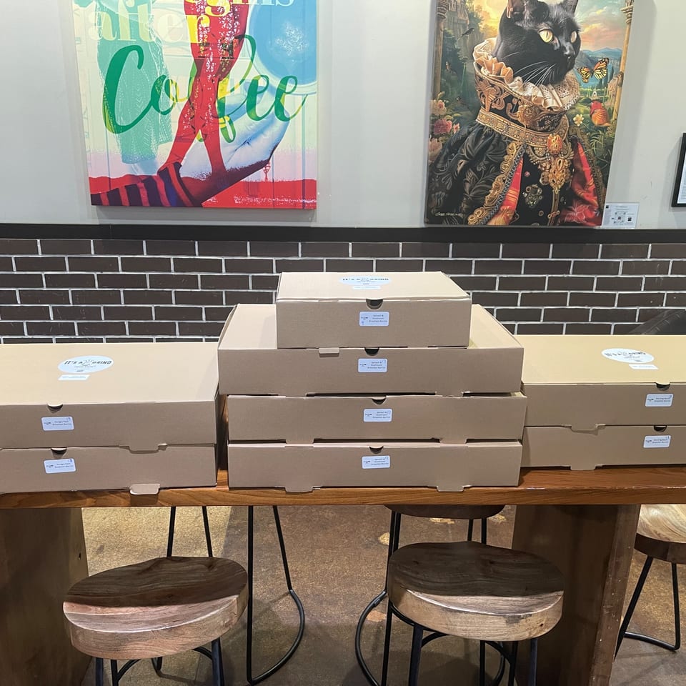 Numerous "It's a Grind" coffee boxes stacked on a wooden table with stools, under vibrant art.