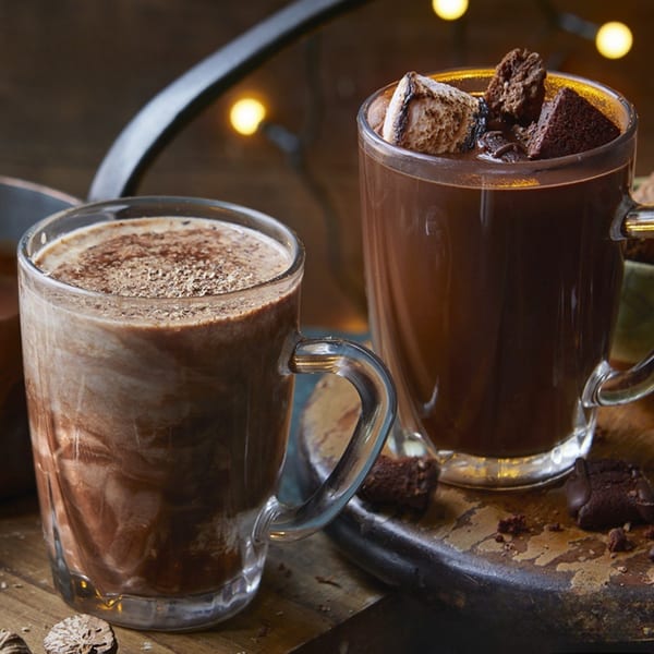 Two mugs of hot chocolate, one with marshmallows and brownie pieces, on a wooden surface.
