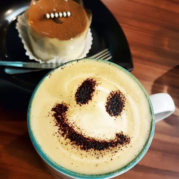 A latte with a smiley face in foam and a small cake on a black plate.