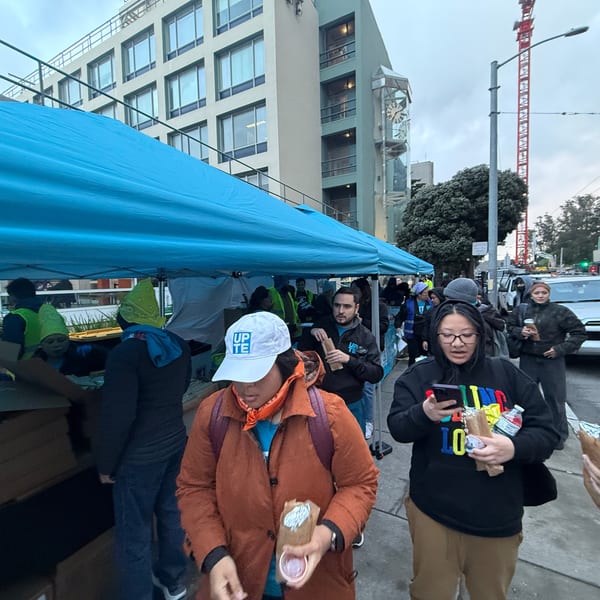 People gather under a blue tent on a city street, some holding food items.