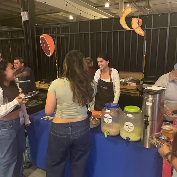 A vendor smiles behind a table with drink dispensers, serving customers at an indoor event.