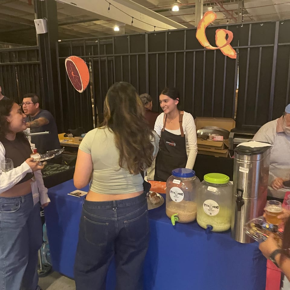 A vendor smiles behind a table with drink dispensers, serving customers at an indoor event.